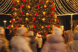 People gathered around a large Christmas tree decorated with golden lights and red ornaments in an Italian piazza at night.