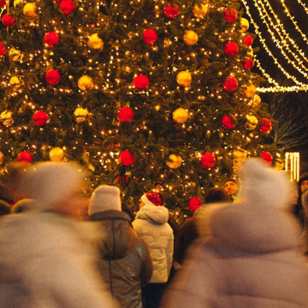People gathered around a large Christmas tree decorated with golden lights and red ornaments in an Italian piazza at night.