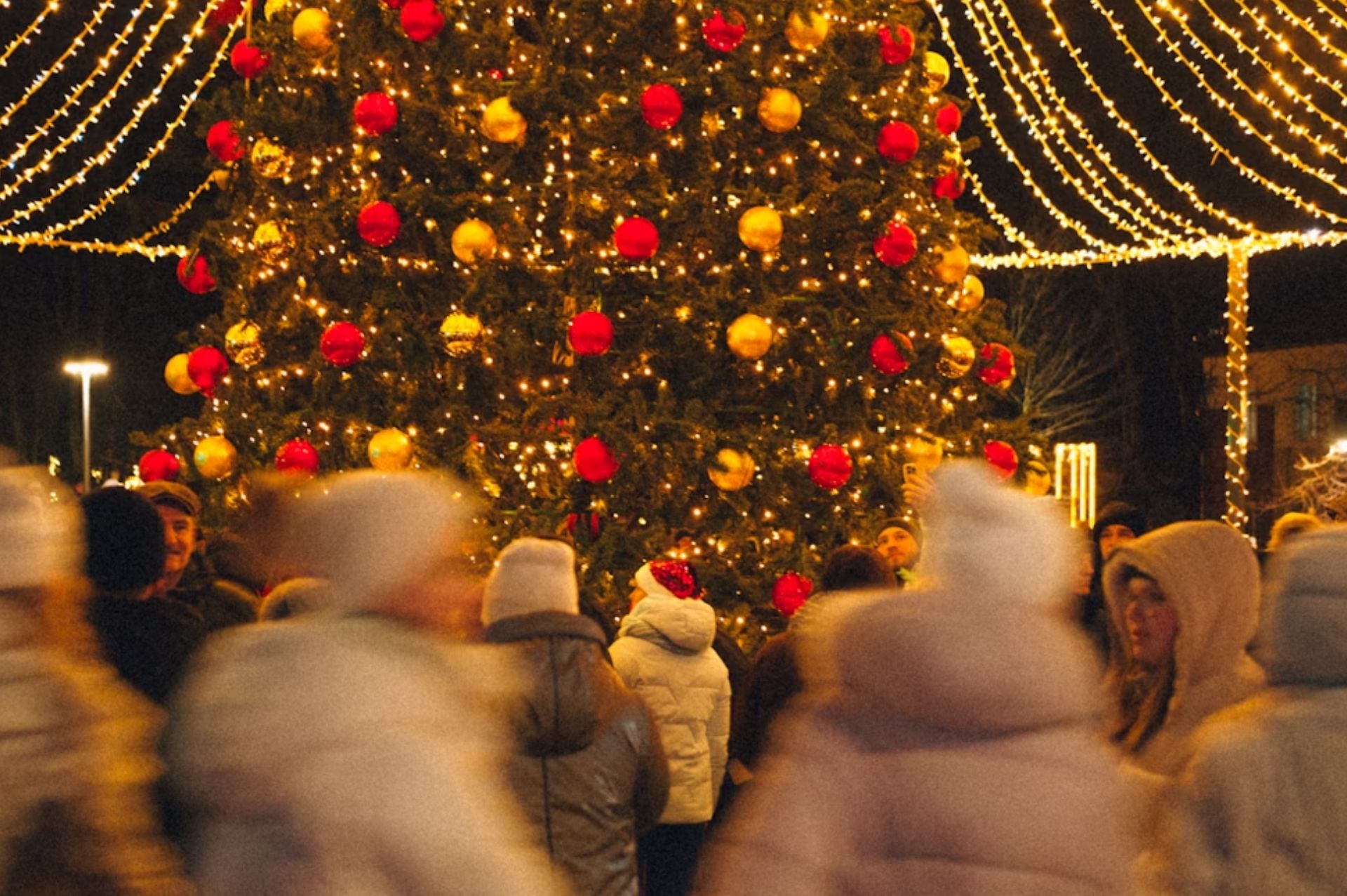 People gathered around a large Christmas tree decorated with golden lights and red ornaments in an Italian piazza at night.