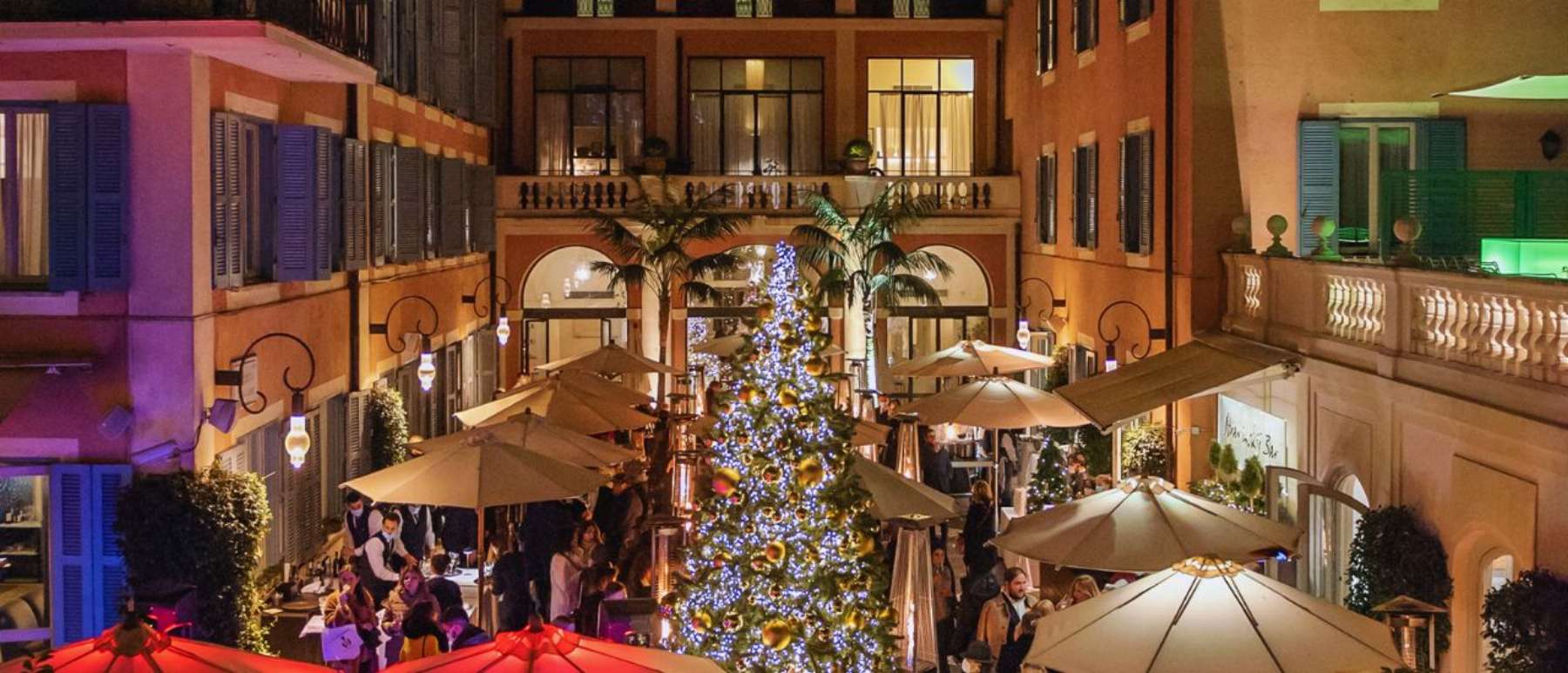 Festive outdoor courtyard with a large decorated Christmas tree, evening lights, and people dining under umbrellas.