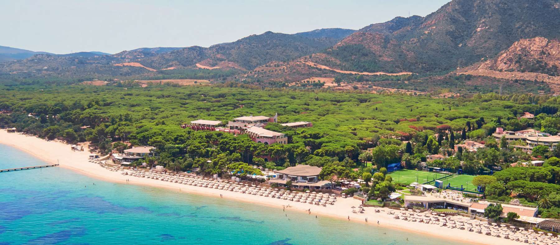 Beach side location of Forte Village Sardinia showing green hills in the background, white sand beach with umbrellas and watersports next to turquoise sea